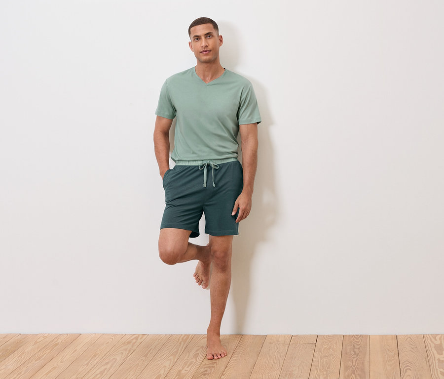 Mężczyzna stoi na jednej nodze oparty o ścianę, ubrany w zielony t-shirt i szorty.