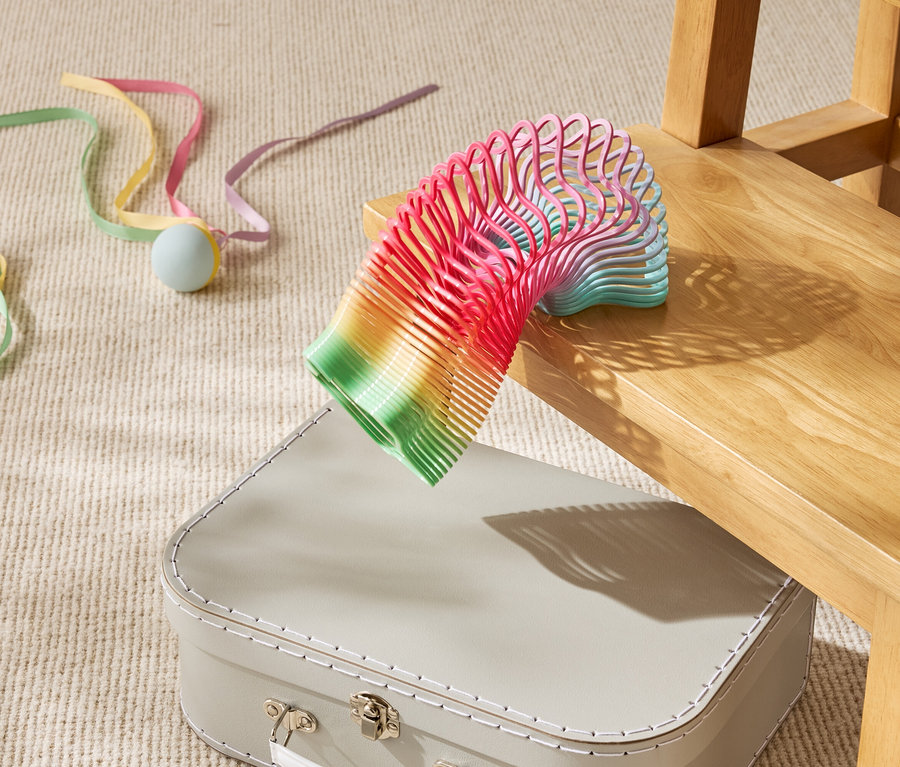 Kolorowa sprężyna-zabawka leży częściowo na beżowej walizce pod drewnianym krzesłem.