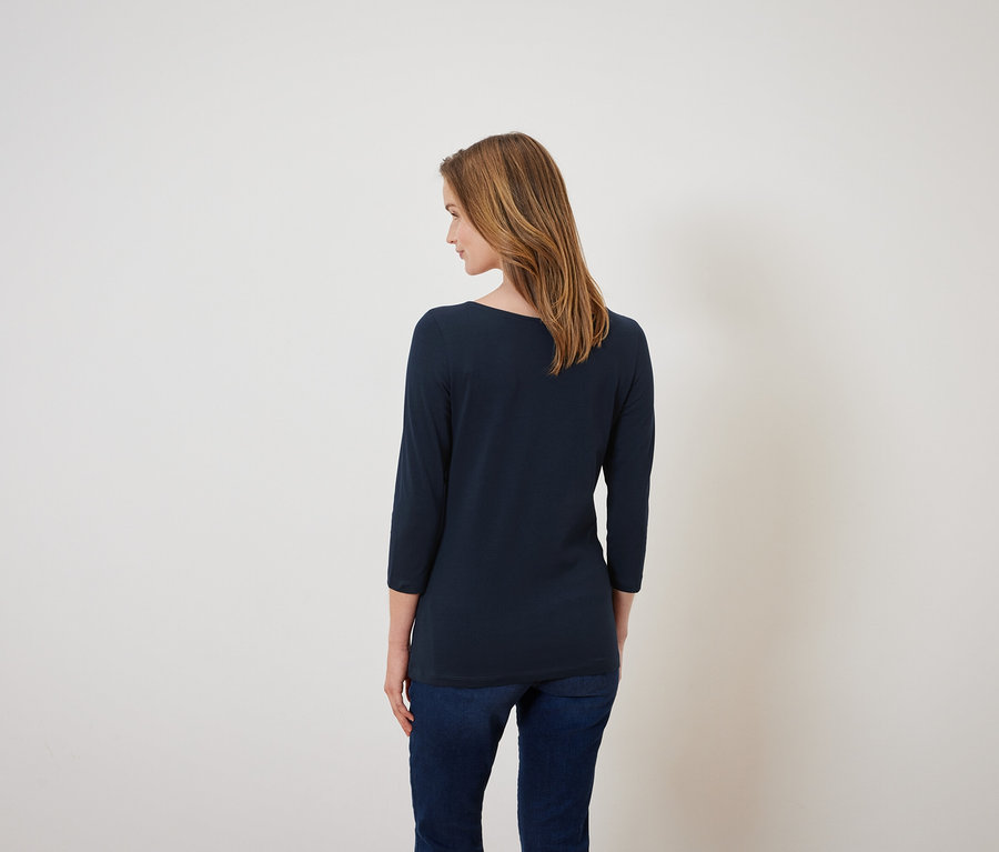 Kobieta stoi tyłem, ubrana w granatową koszulkę z rękawem trzy czwarte i dżinsy.