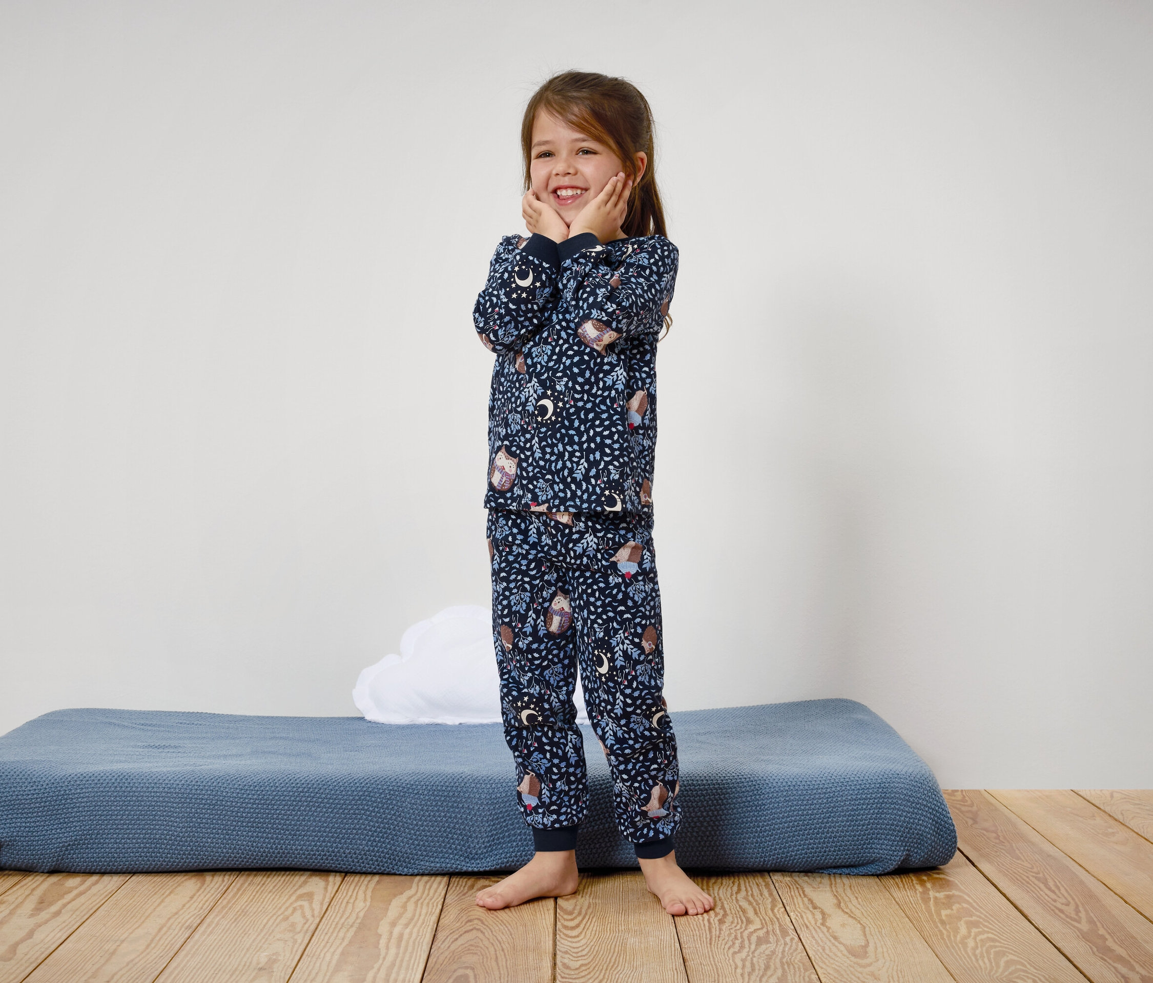 Uśmiechnięta dziewczynka w niebieskiej piżamie w nadruki stoi na drewnianej podłodze.