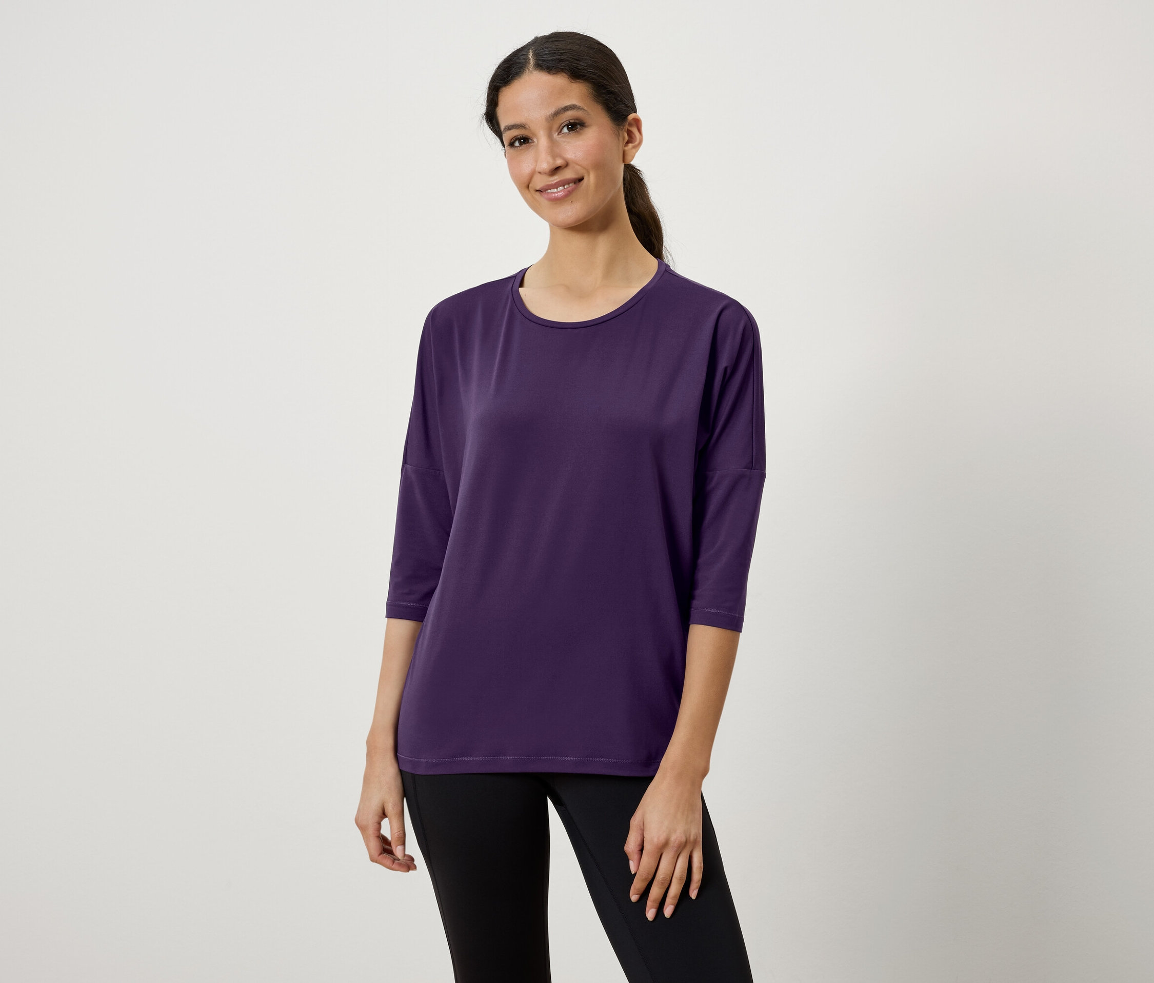 Kobieta o brązowych włosach związanych w kucyk ma na sobie fioletową koszulkę z rękawami trzy czwarte i czarne legginsy.
