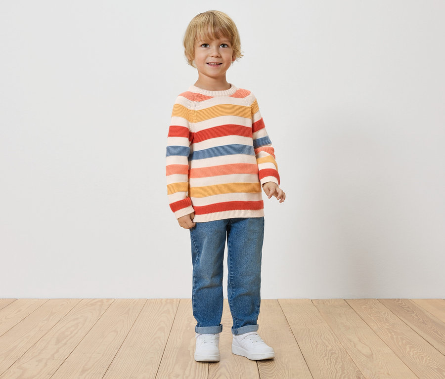 Chłopiec z blond włosami stoi na tle białej ściany, ubrany w pasiasty sweter, dżinsy i białe tenisówki.