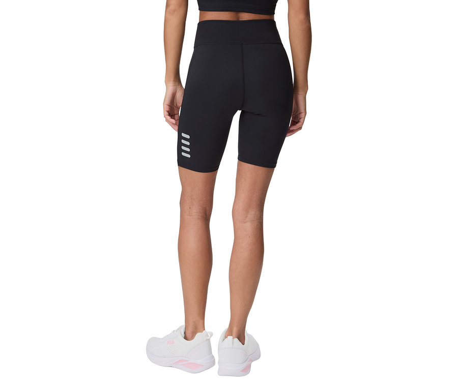 Kobieta od tyłu w czarnych spodenkach rowerowych z czterema odblaskowymi paskami i białych butach sportowych.