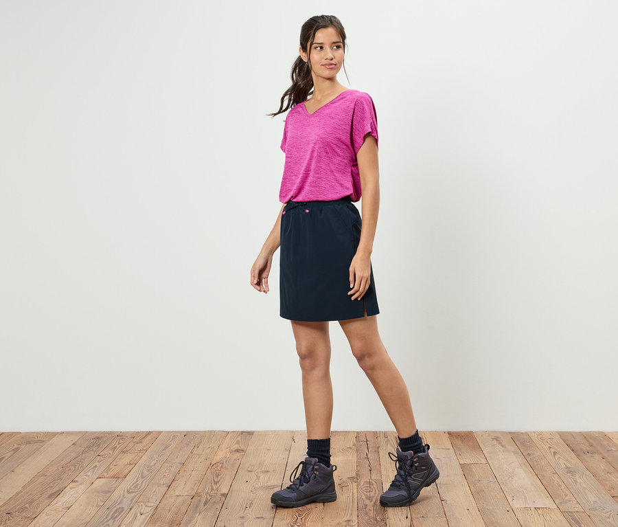 Kobieta stoi na tle białej ściany, ubrana w różową koszulkę, czarną spódnicę i buty trekkingowe.