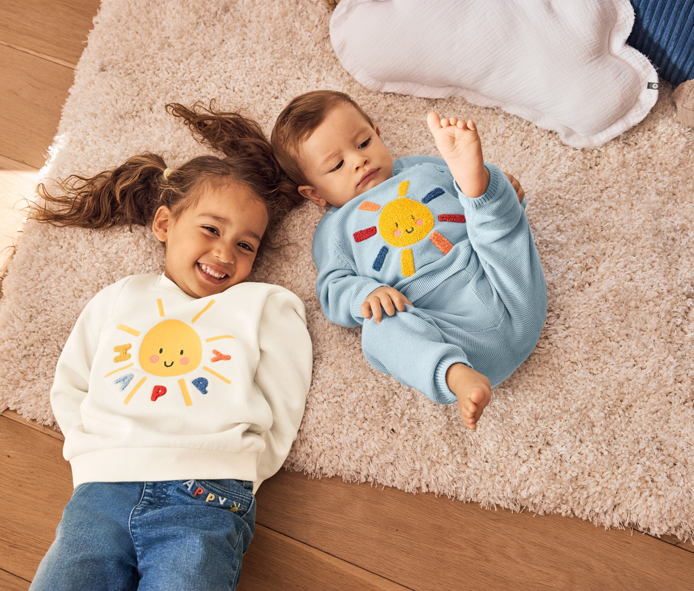 Dziewczynka z warkoczami i niemowlę leżą na dywanie. Dziewczynka ma na sobie kremową bluzę z żółtym słońcem. Niemowlę ma na sobie niebieski kombinezon z dzianiny ze słońcem.
