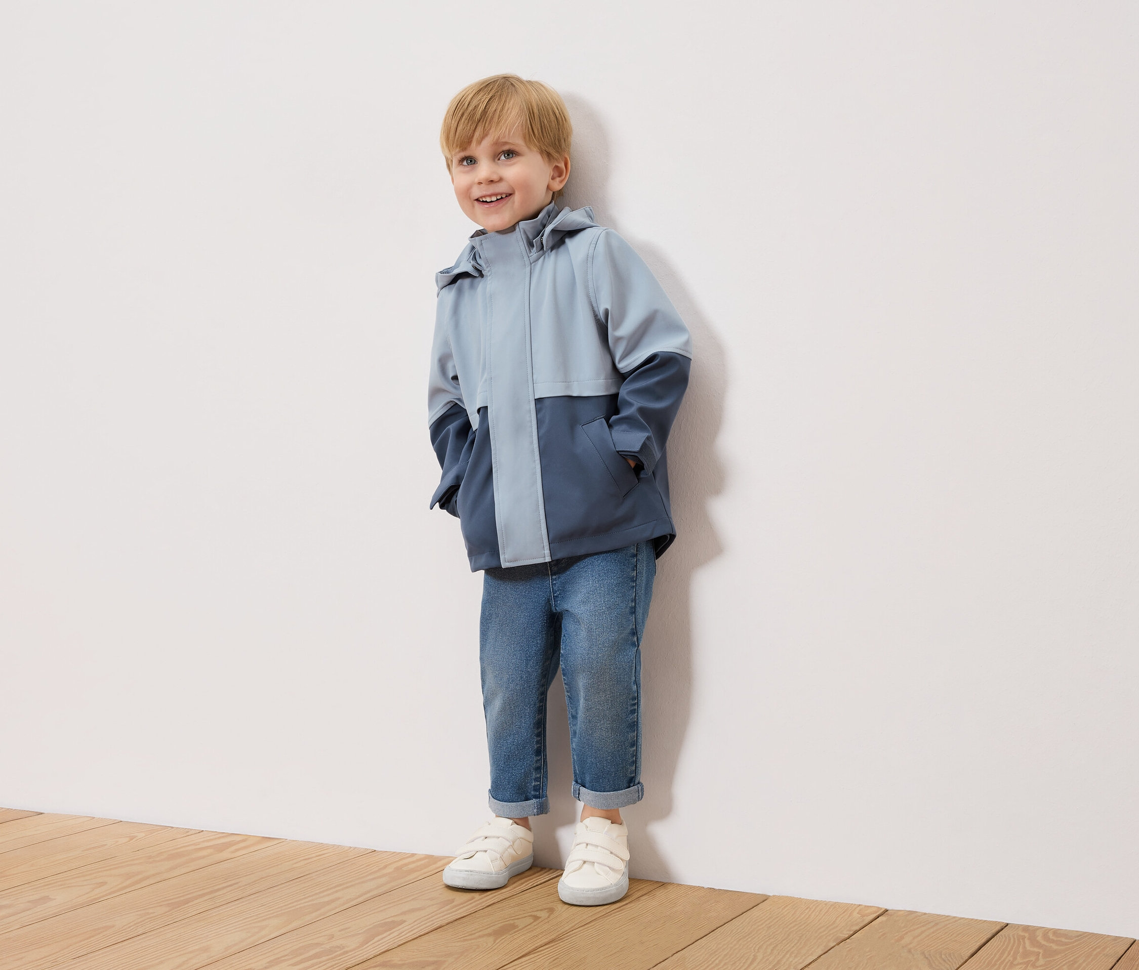 Uśmiechnięty blondwłosy chłopiec stoi na tle białej ściany, ubrany w dwukolorową, błękitno-granatową kurtkę, niebieskie dżinsy i białe tenisówki.