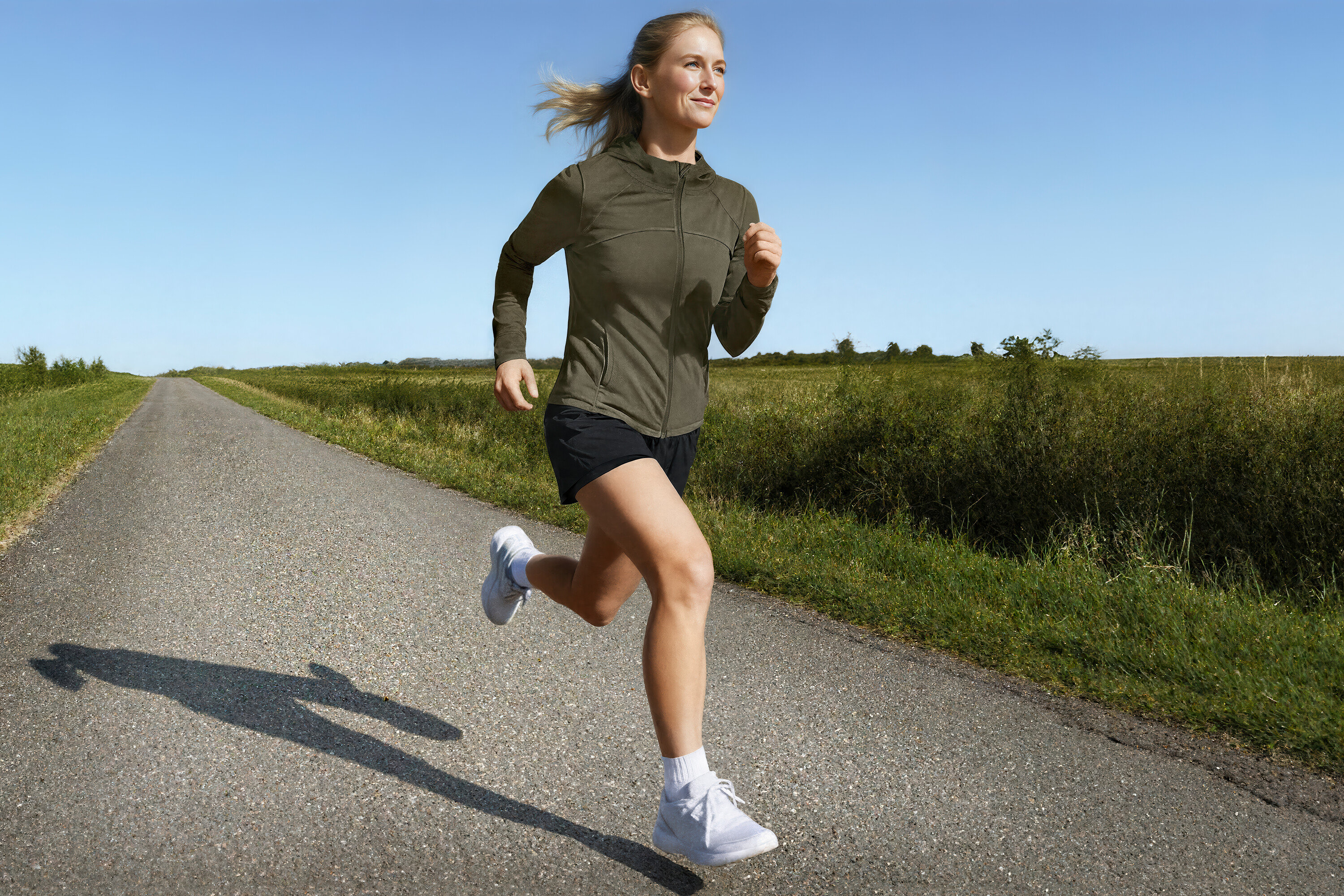 Kobieta biegnie w kurtce sportowej i szortach do biegania 2 w 1 po wiejskiej drodze.