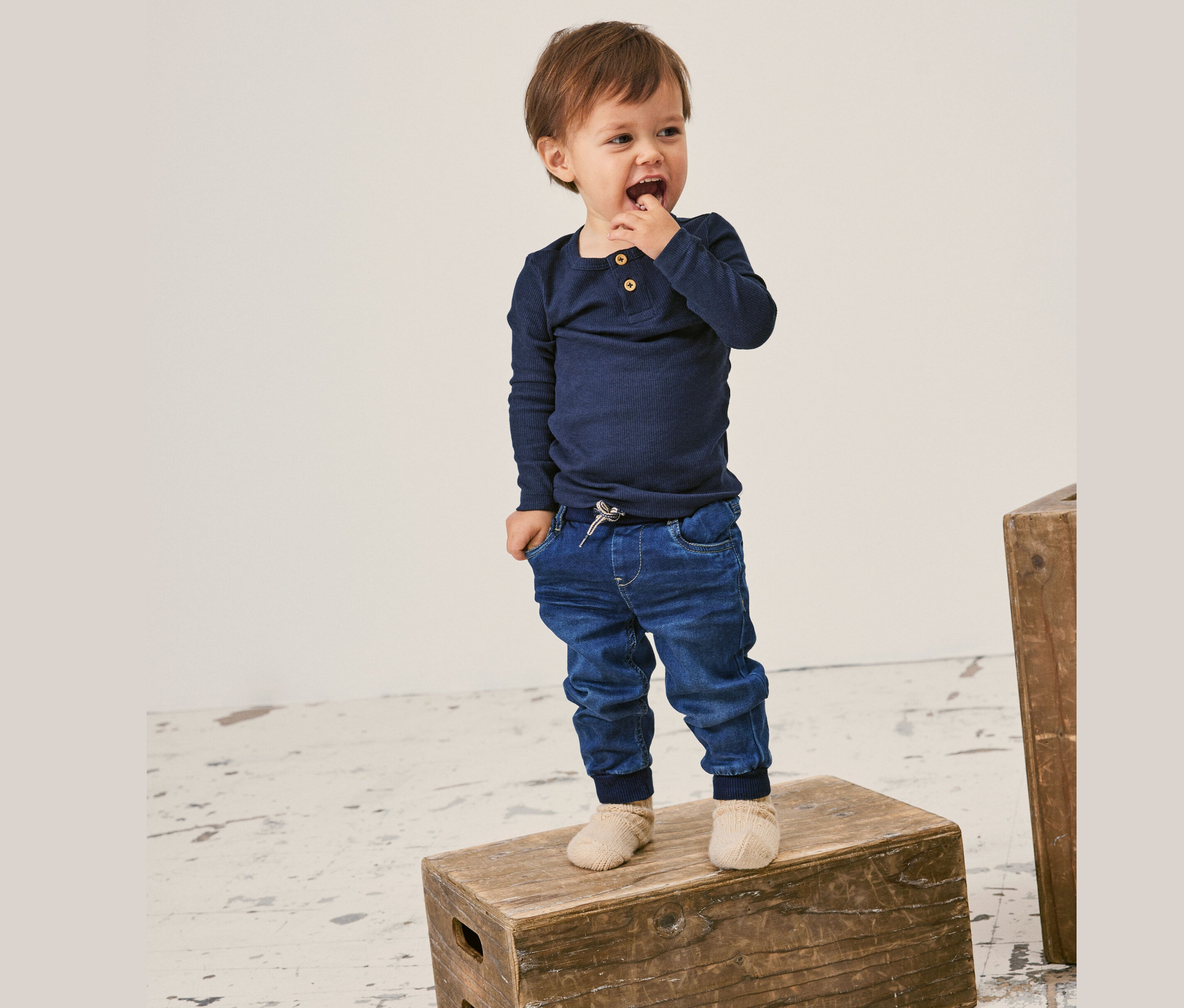 Mały chłopiec z brązowymi włosami, ubrany w granatową koszulkę z długim rękawem, dżinsy i skarpetki, stoi na drewnianej skrzyni.