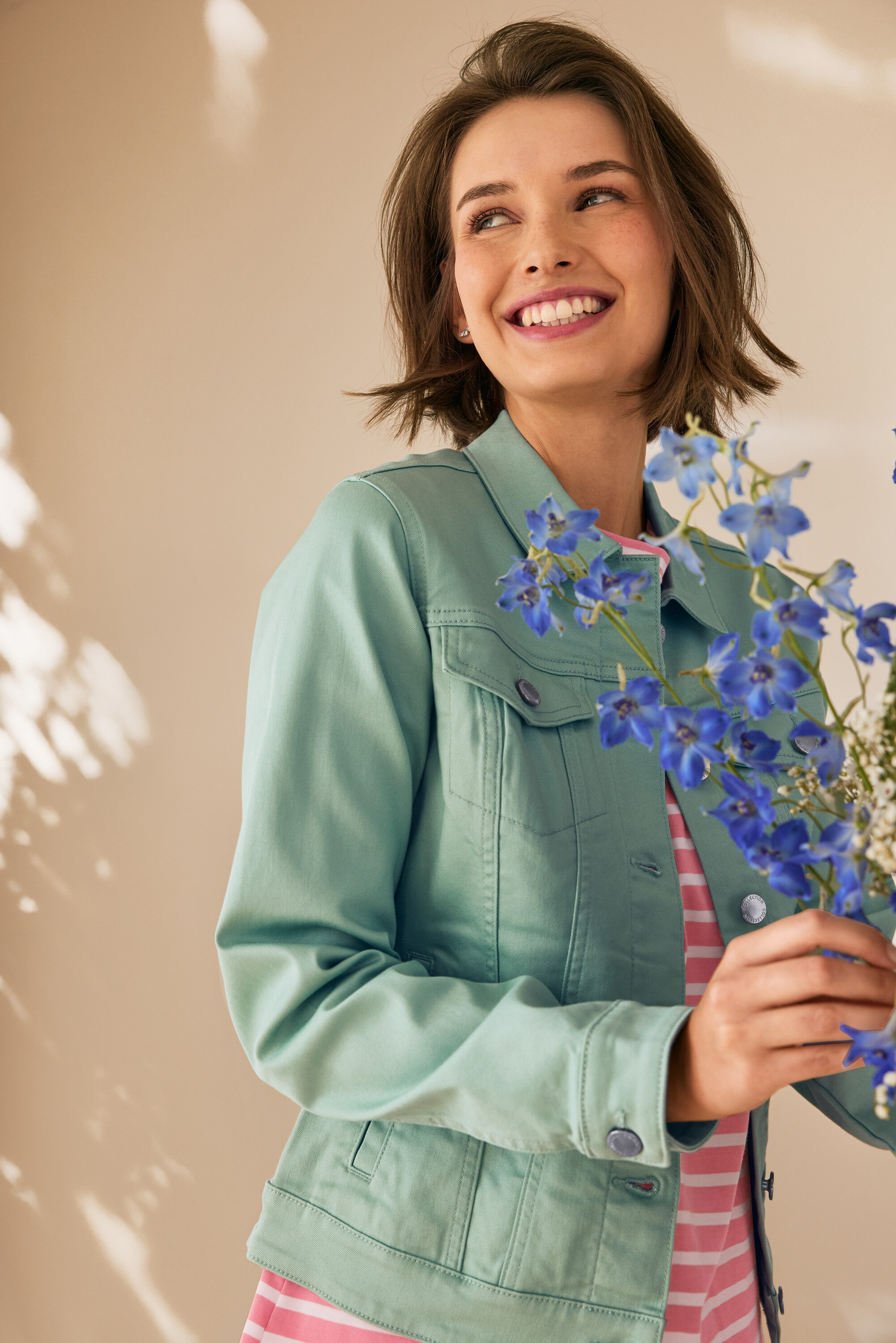 Uśmiechnięta kobieta trzyma niebieskie kwiaty, ma na sobie miętową kurtkę dżinsową.