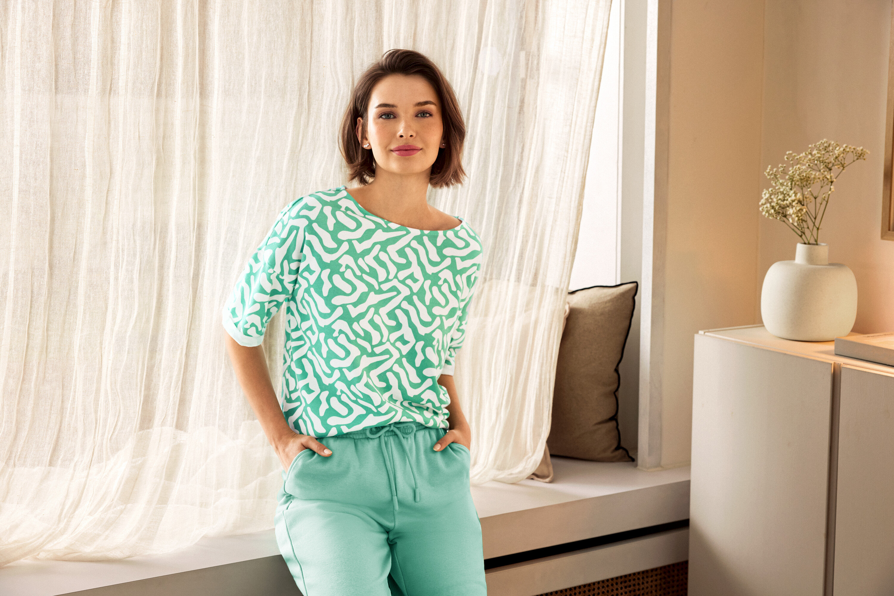 Kobieta w zielonych spodniach i wzorzystej bluzce pozuje obok okna. W tle widać wazon.