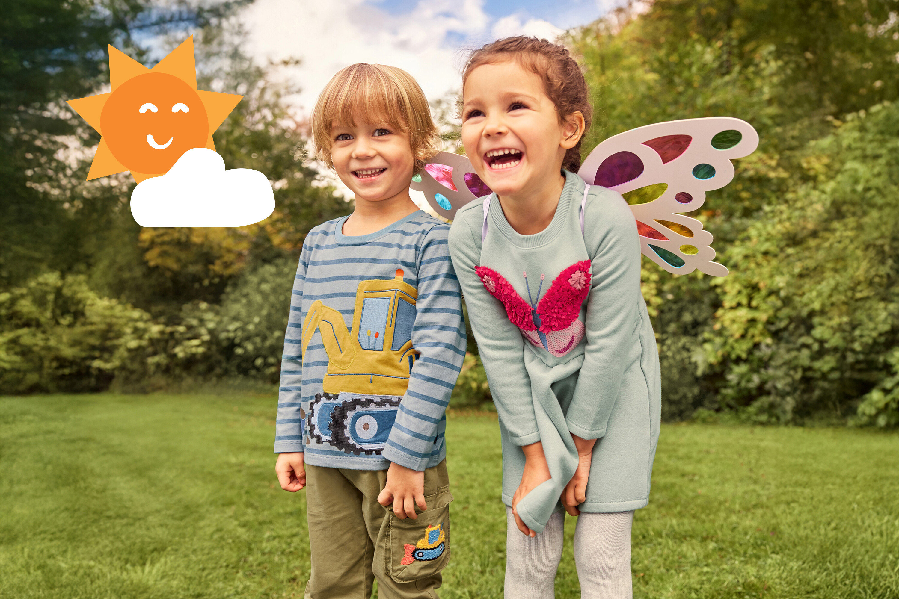 Dwoje roześmianych dzieci stoi na trawie. Dziewczynka ma na sobie skrzydła i sukienkę z motylem, a chłopiec koszulkę z koparką.