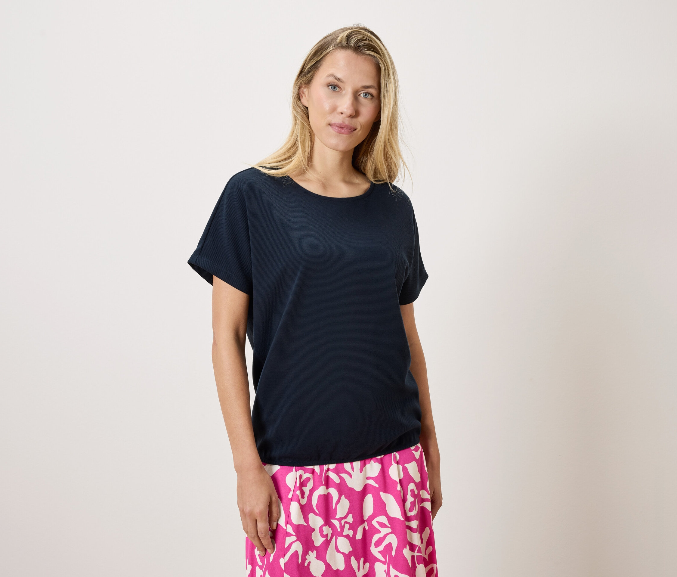 Blondwłosa kobieta ma na sobie granatową koszulkę i różowo-białą spódnicę na białym tle.