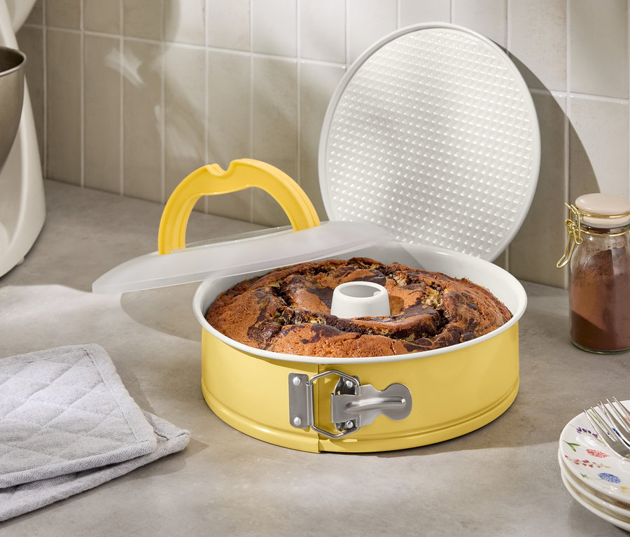 Ciasto marmurkowe w żółtej formie do pieczenia z pokrywką i uchwytem obok robota kuchennego i słoika z przyprawami