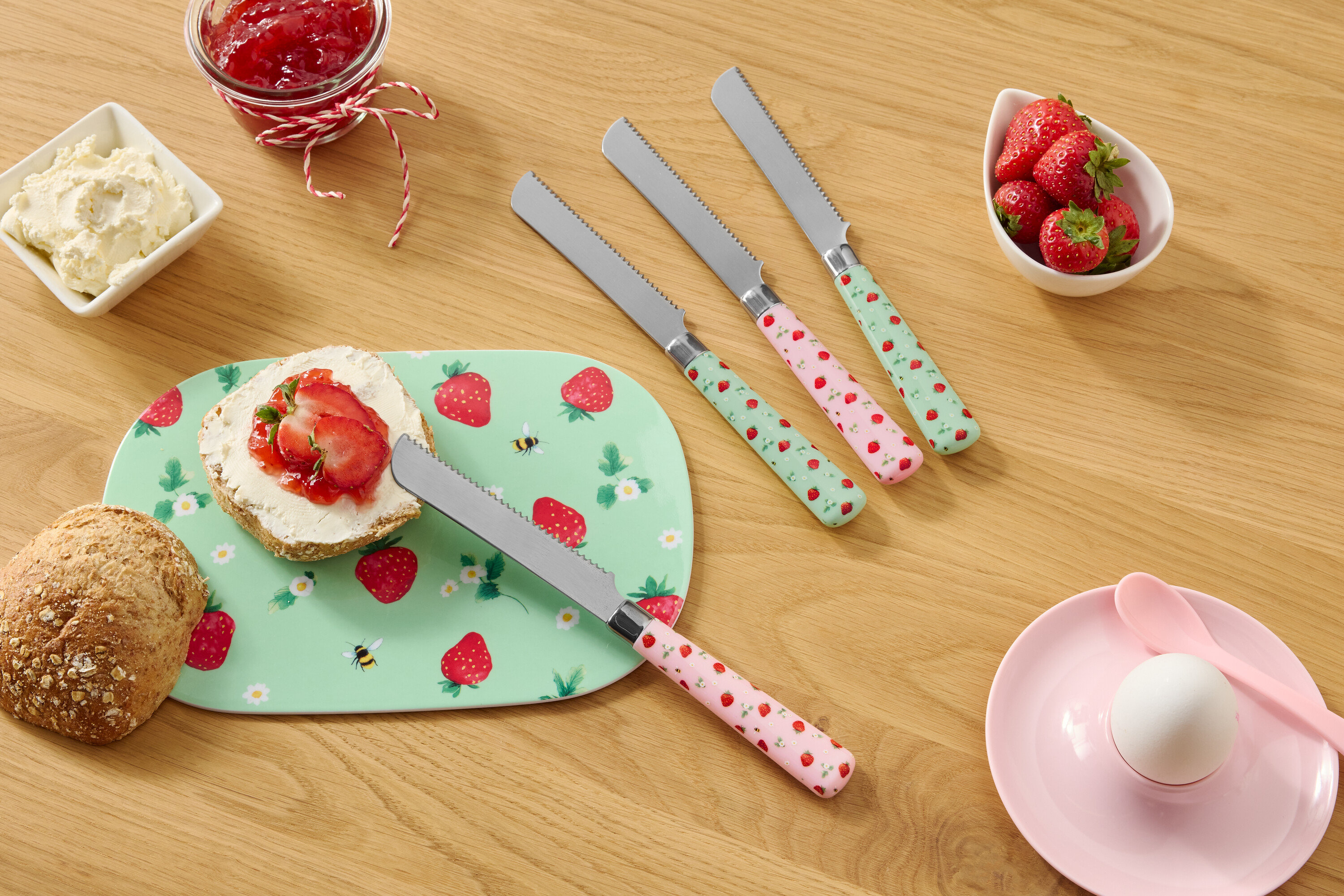 Na drewnianym stole znajdują się bułka, talerz we wzór truskawek, serek śmietankowy, dżem, truskawki i cztery noże we wzór truskawek.