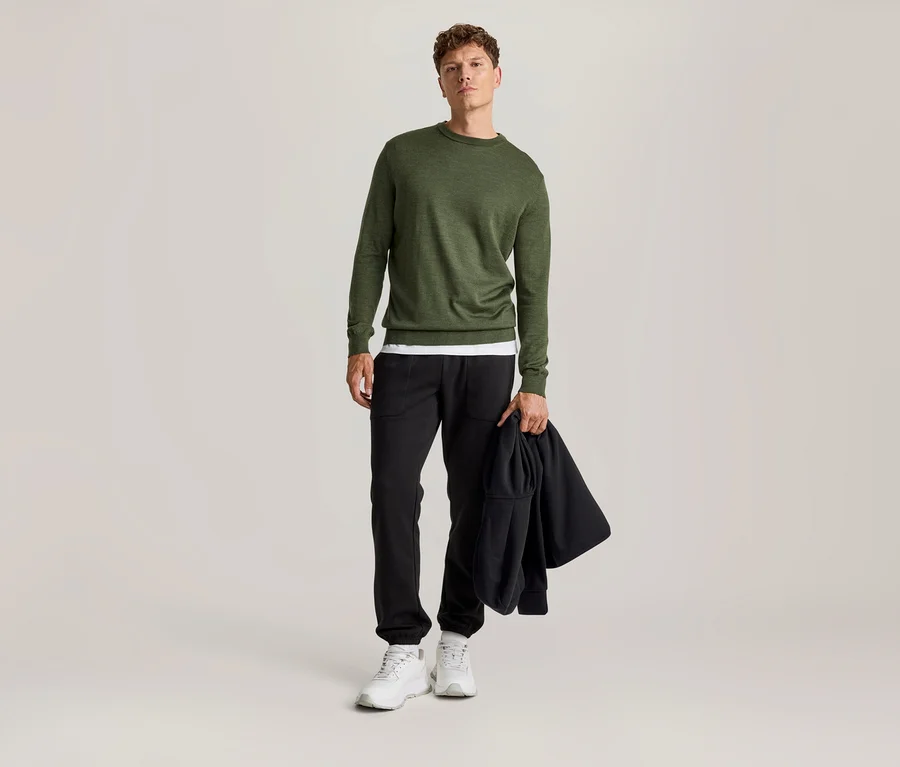 Mężczyzna stoi, ubrany w zielony sweter z długim rękawem na białym T-shircie, czarne spodnie i białe sneakersy. W ręku trzyma czarną kurtkę.