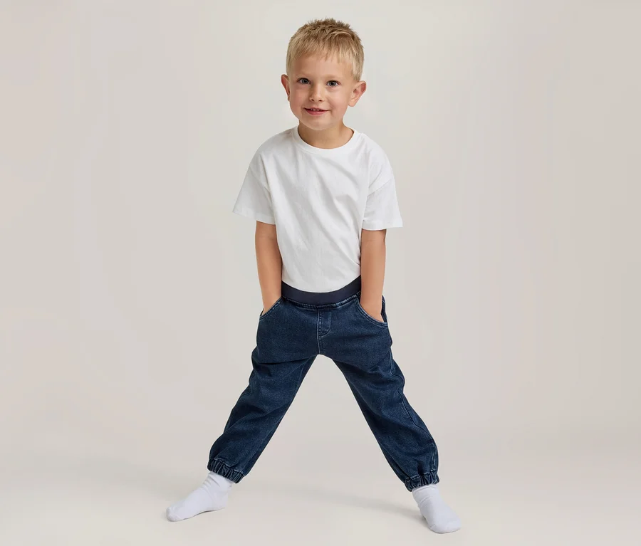 Młody chłopiec z blond włosami uśmiecha się, ubrany w biały T-shirt i ciemnoniebieskie spodnie joggery z rękami w kieszeniach, stoi w rozkroku w białych skarpetkach.