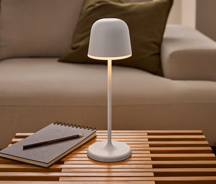 Świecąca biała akumulatorowa lampa stołowa LED Eglo „Mannera” stoi na drewnianym stole lamelowym obok notatnika z ołówkiem, przed sofą.
