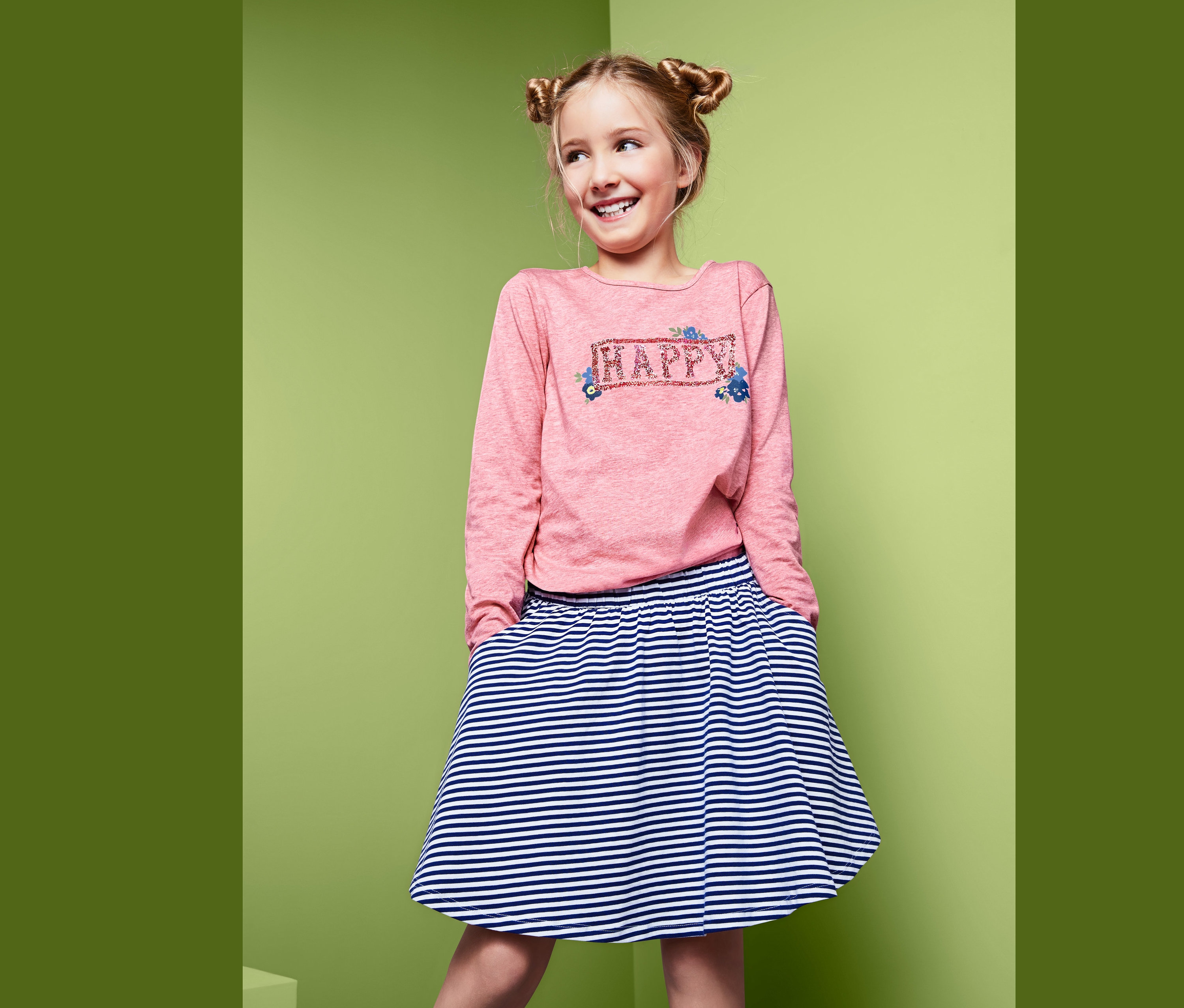 Uśmiechnięta dziewczyna pozuje w różowej bluzce z długim rękawem i dwustronnymi cekinami oraz w spódnicy w paski.