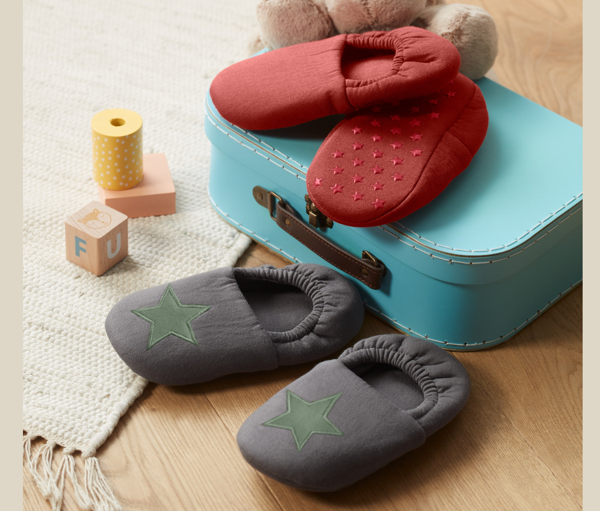 Dwie pary kapci dla małych dzieci leżą na niebieskiej walizce, jedna para czerwona w gwiazdki, druga szara z zieloną gwiazdą.