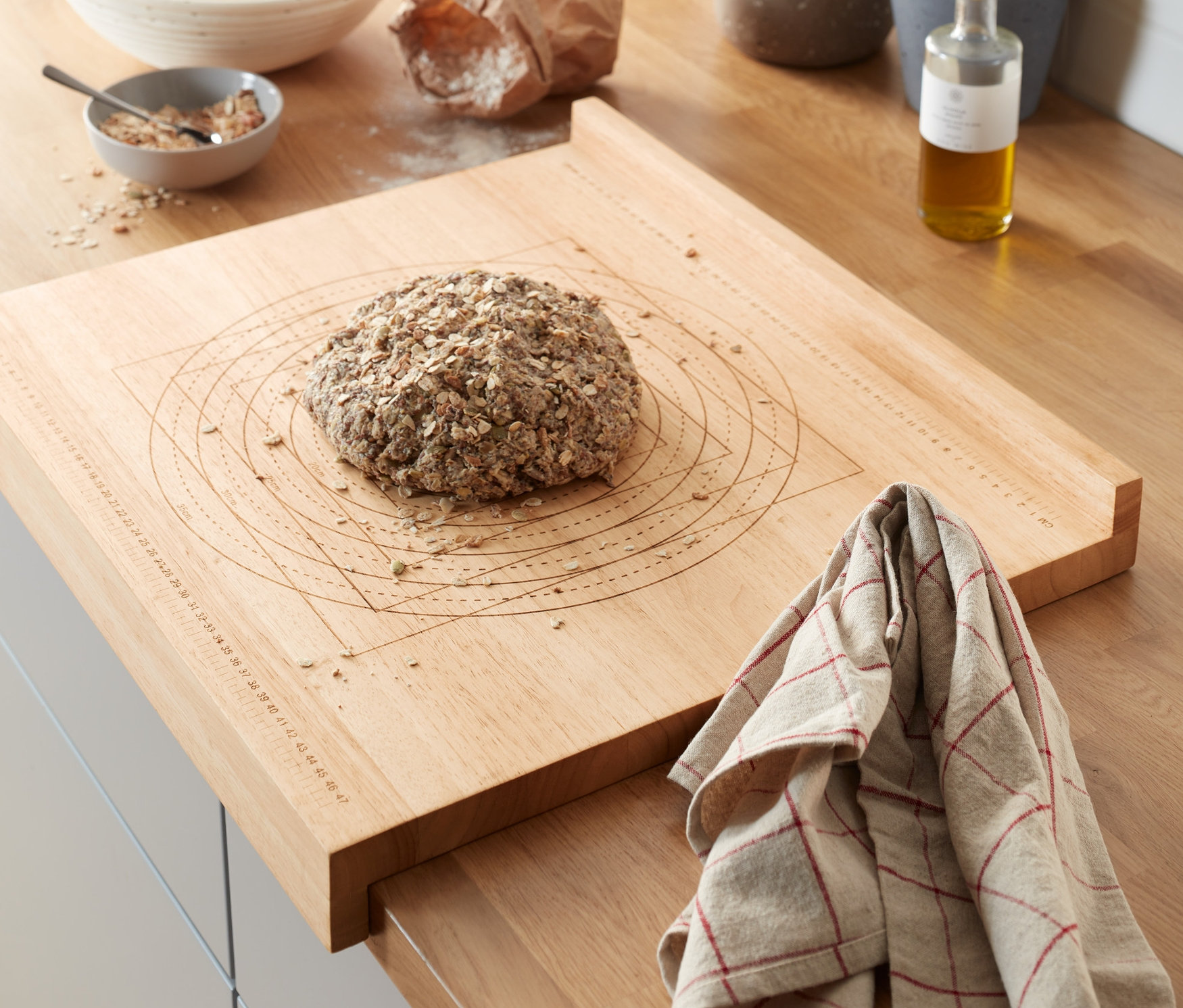 Na blacie kuchennym leży drewniana deska do prac kuchennych i przygotowywania wypieków z bochenkiem chleba. W tle zioła w doniczkach i inne przybory kuchenne.