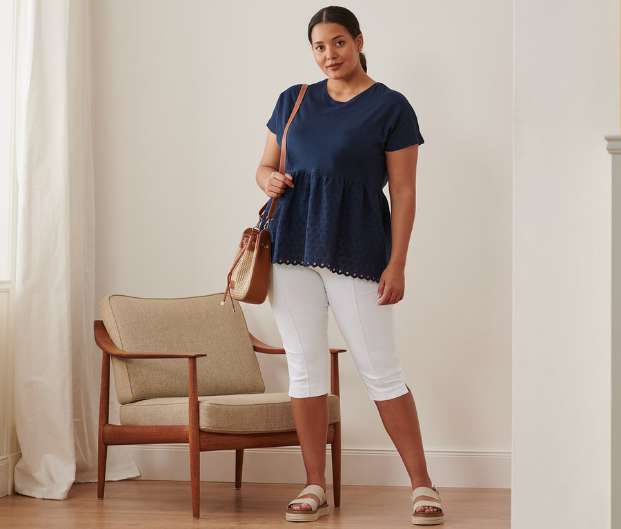 Kobieta stoi, ubrana w granatową bluzkę z mieszanki materiałowej z haftem, spodnie stretchowe o długości 3/4, sandały skórzane na platformie i torbę-worek.