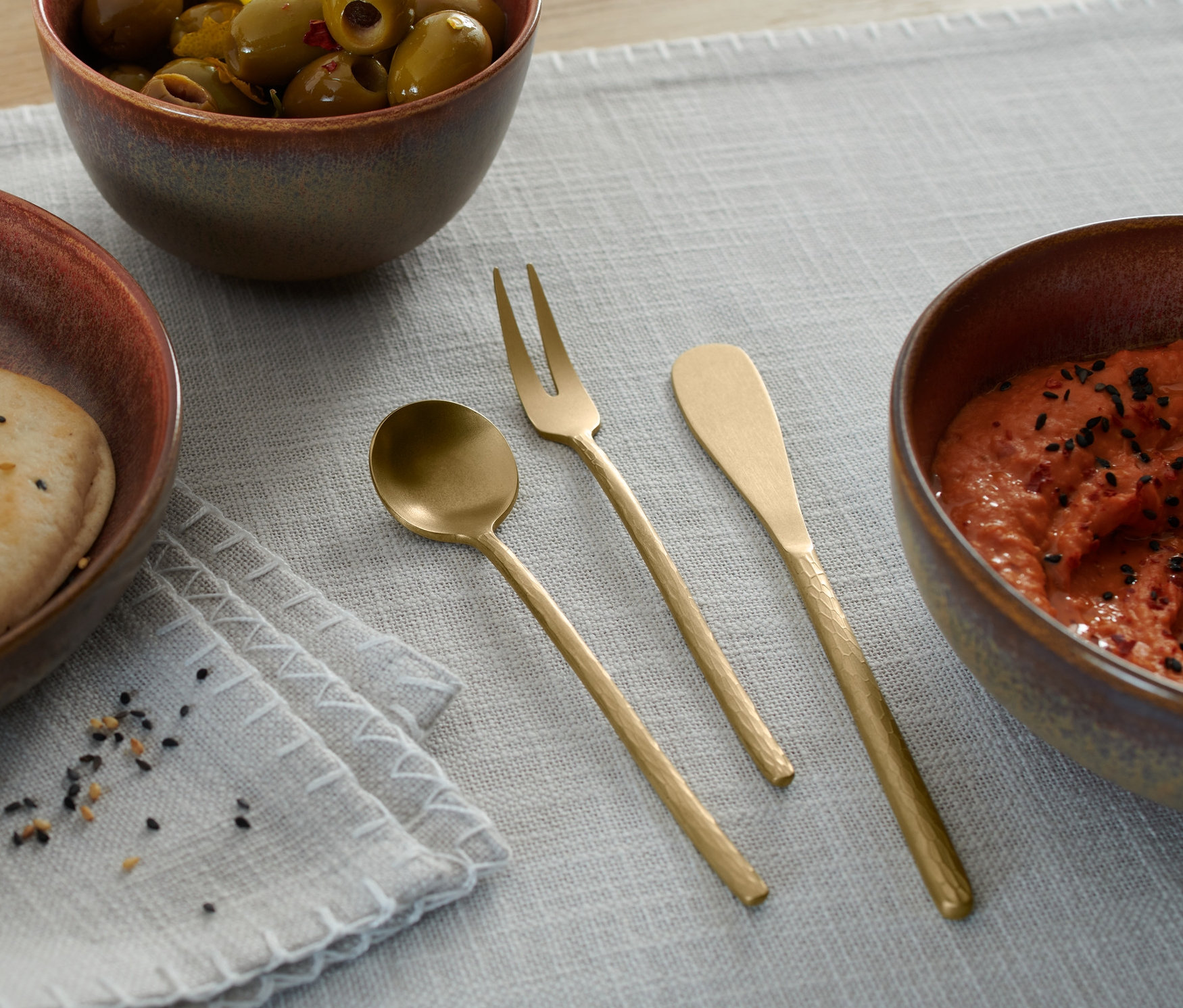 Na stole leżą miska z chlebem, miska z oliwkami, miska z dipem oraz zestaw sztućców (3 sztuki).