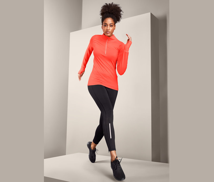 Kobieta pozuje w neonowo-pomarańczowej koszulce funkcyjnej i czarnych legginsach.