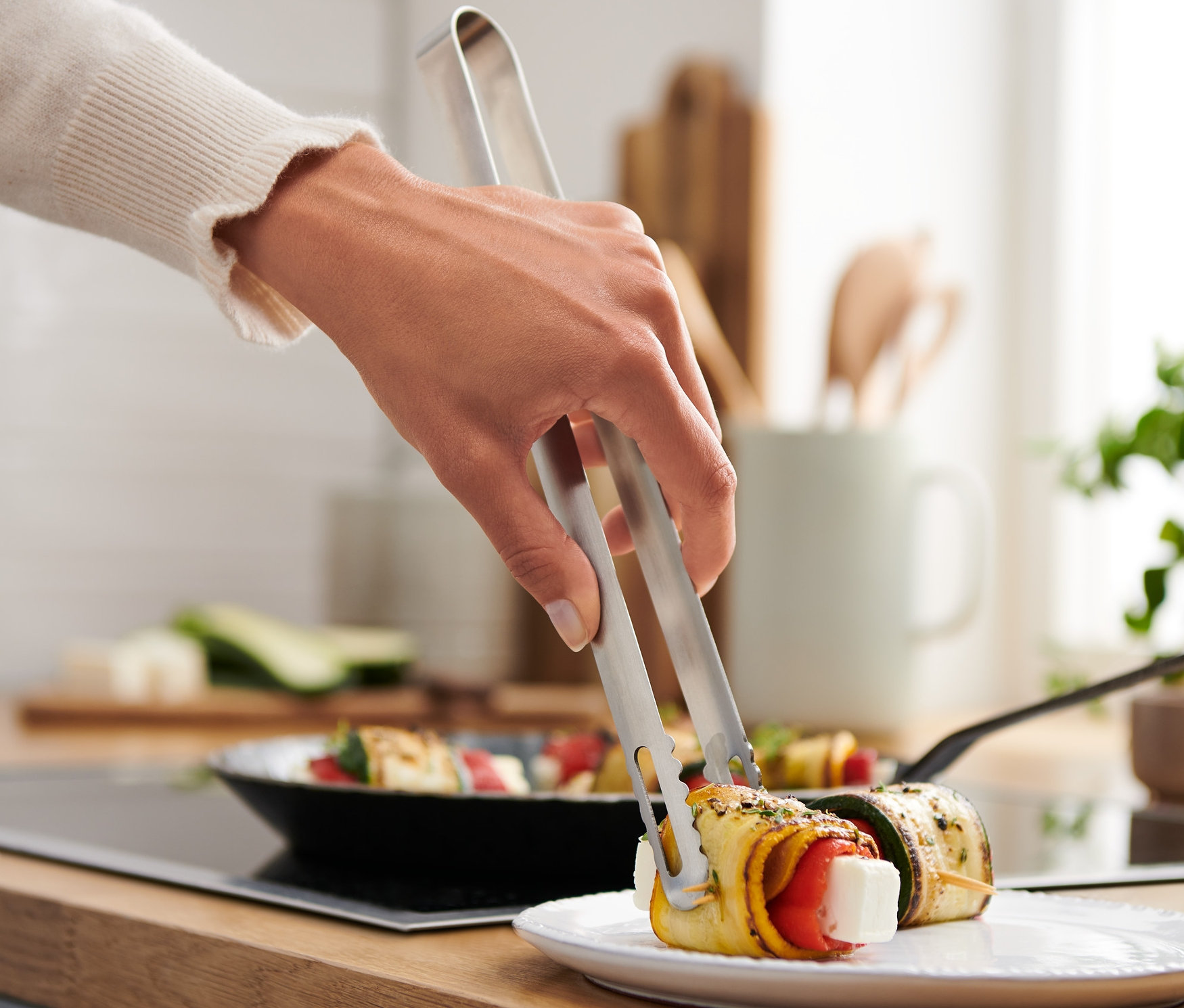 Ręka używa szczypiec kuchennych ze stali nierdzewnej, aby chwycić roladkę z cukinii wypełnioną warzywami na talerzu.