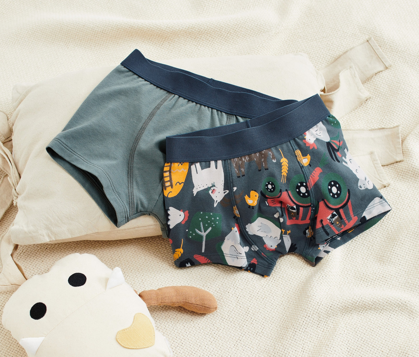 Dwie pary chłopięcych bokserek leżą na beżowej tkaninie obok pluszowej zabawki.