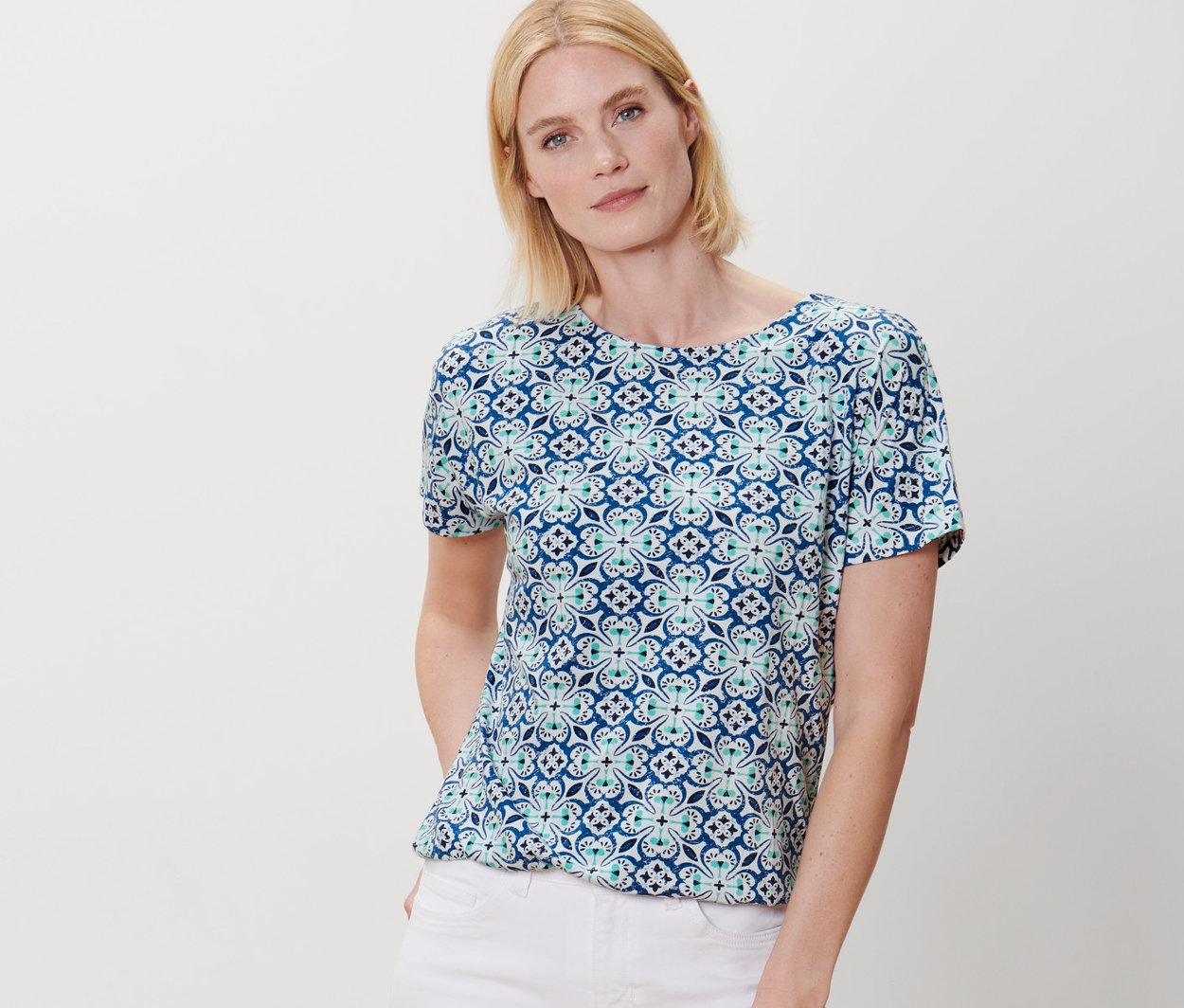 Kobieta pozuje w bluzce z wzorem na całej powierzchni i białych spodniach.