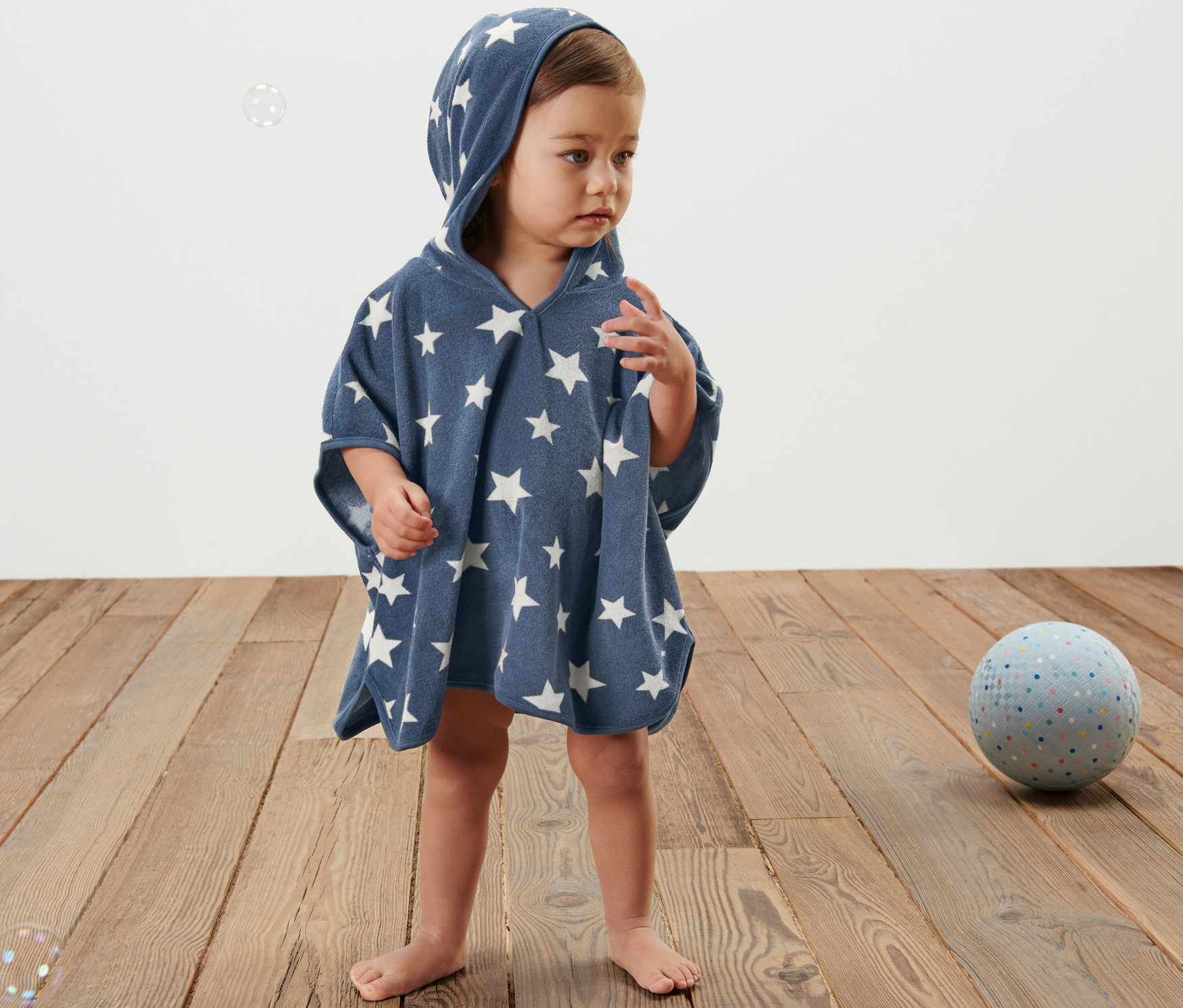 Dziecko stoi na drewnianej podłodze, ubrane w niebieskie ponczo frotte z kapturem i białymi gwiazdami.