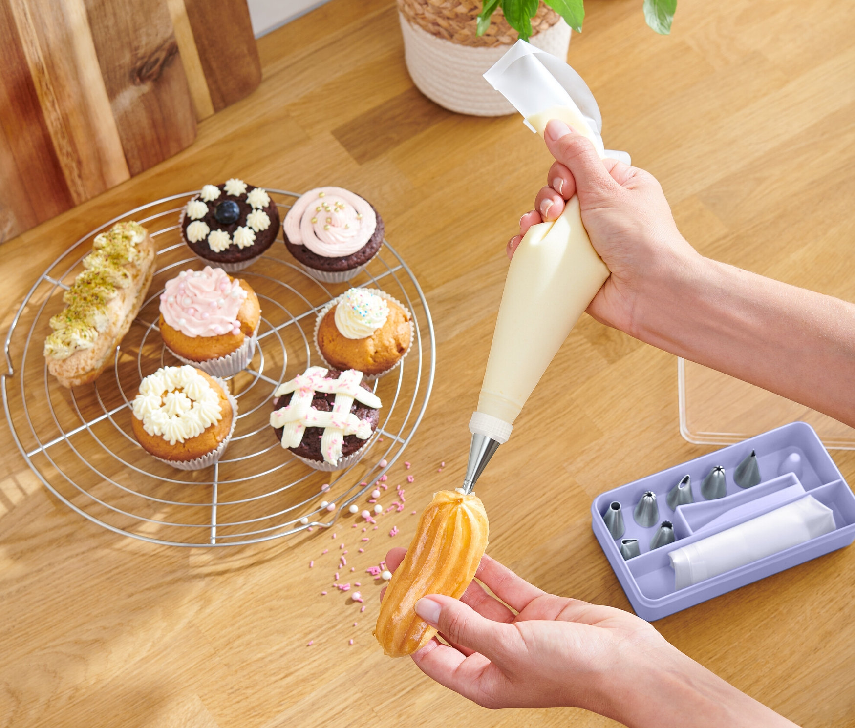 Osoba dekoruje eklerkę kremem za pomocą zestawu do dekorowania ciast. Na stole leżą babeczki i miska z owocami.