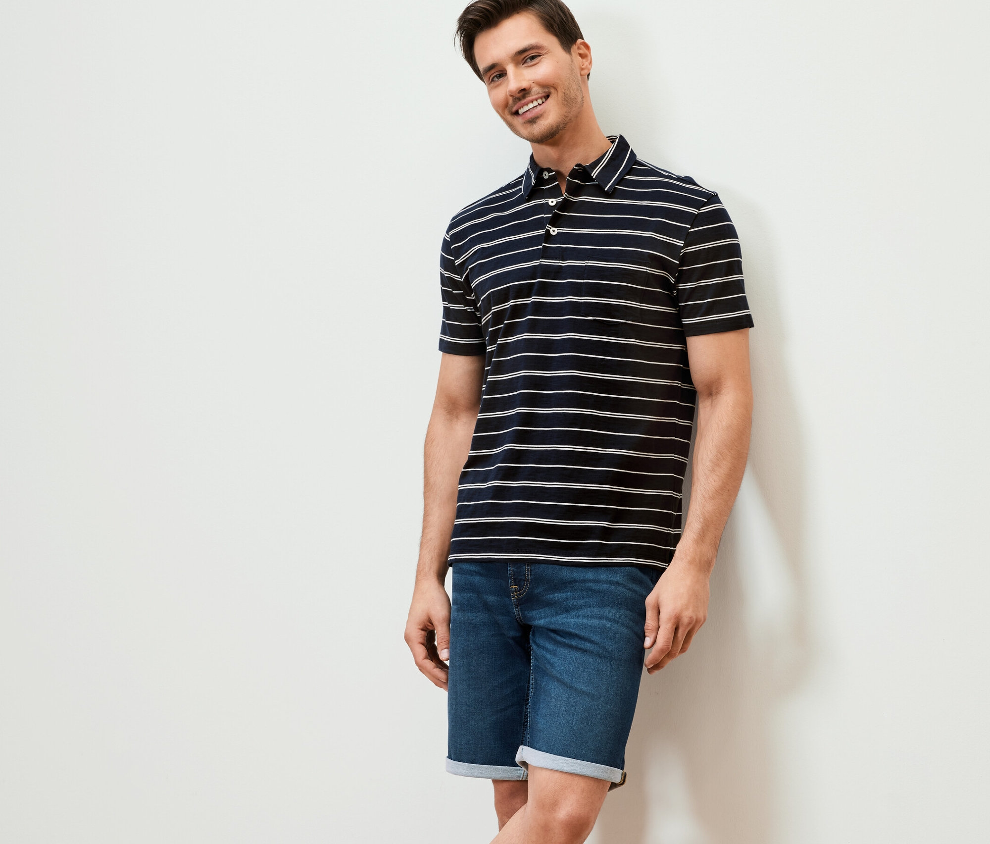 Uśmiechnięty mężczyzna opiera się o ścianę. Ma na sobie granatową w paski dżersejową koszulkę polo i dżinsowe szorty.