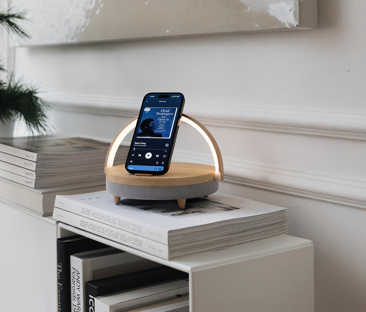 Telefon na głośniku Bluetooth z bezprzewodową ładowarką i lampką LED na stosie książek. Na telefonie widać aplikację muzyczną.
