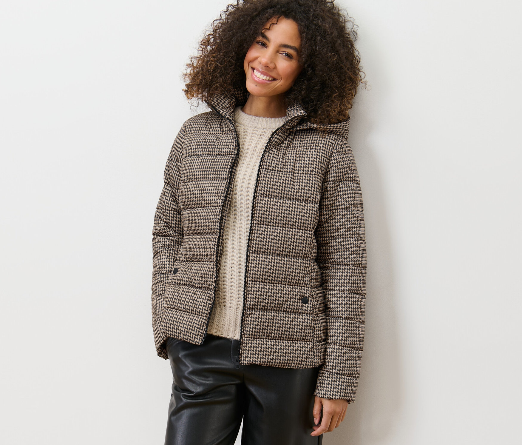 Uśmiechnięta kobieta opiera się o ścianę w pikowanej kurtce z kapturem w pepitkę.