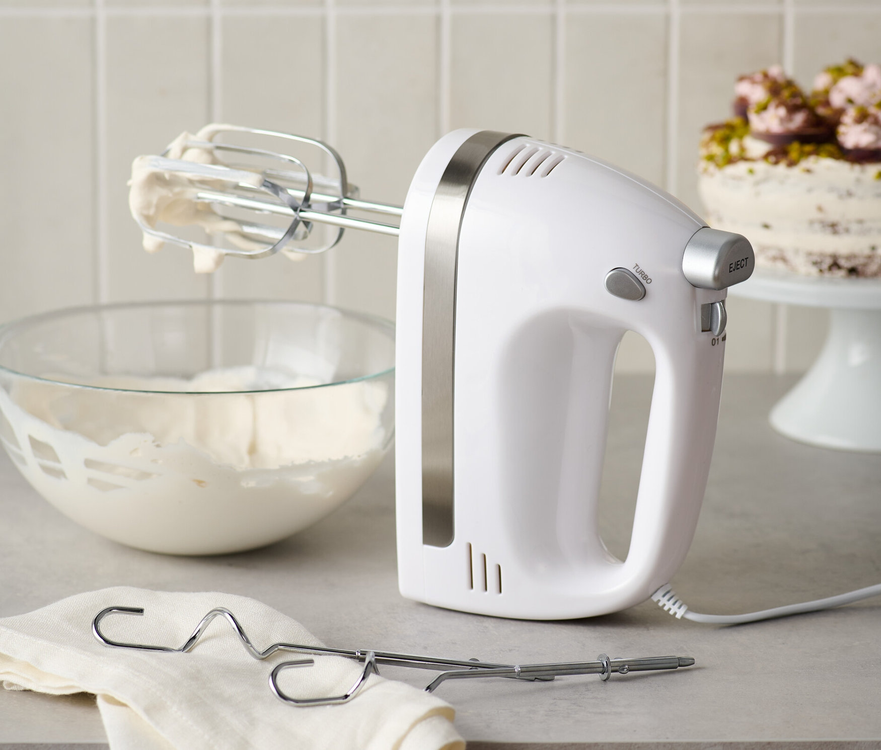 Biały mikser ręczny ze stalowymi mieszadłami stoi obok miski z bitą śmietaną i ściereczki.