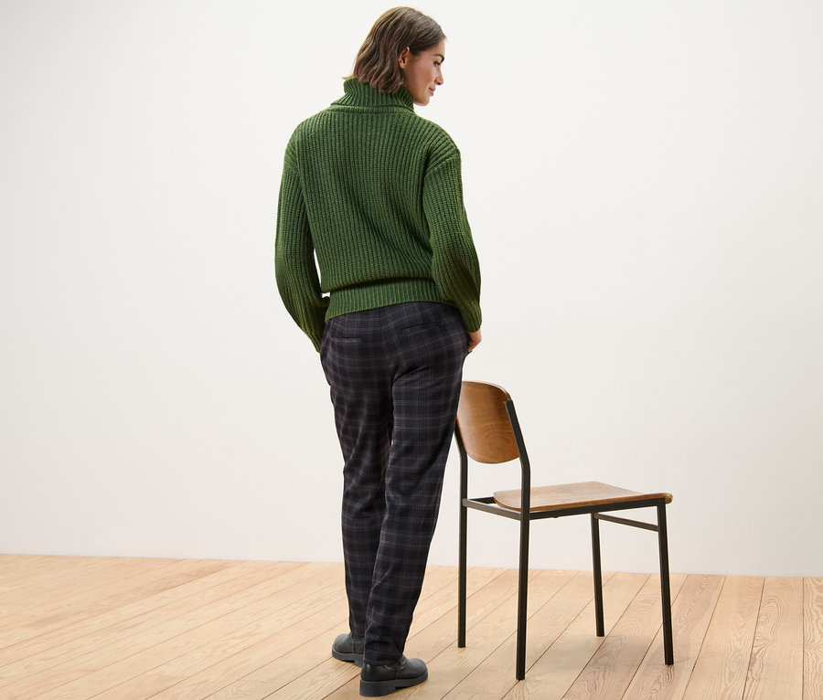 Kobieta stoi tyłem do aparatu, ma na sobie zielony sweter i spodnie joggery w kratę.