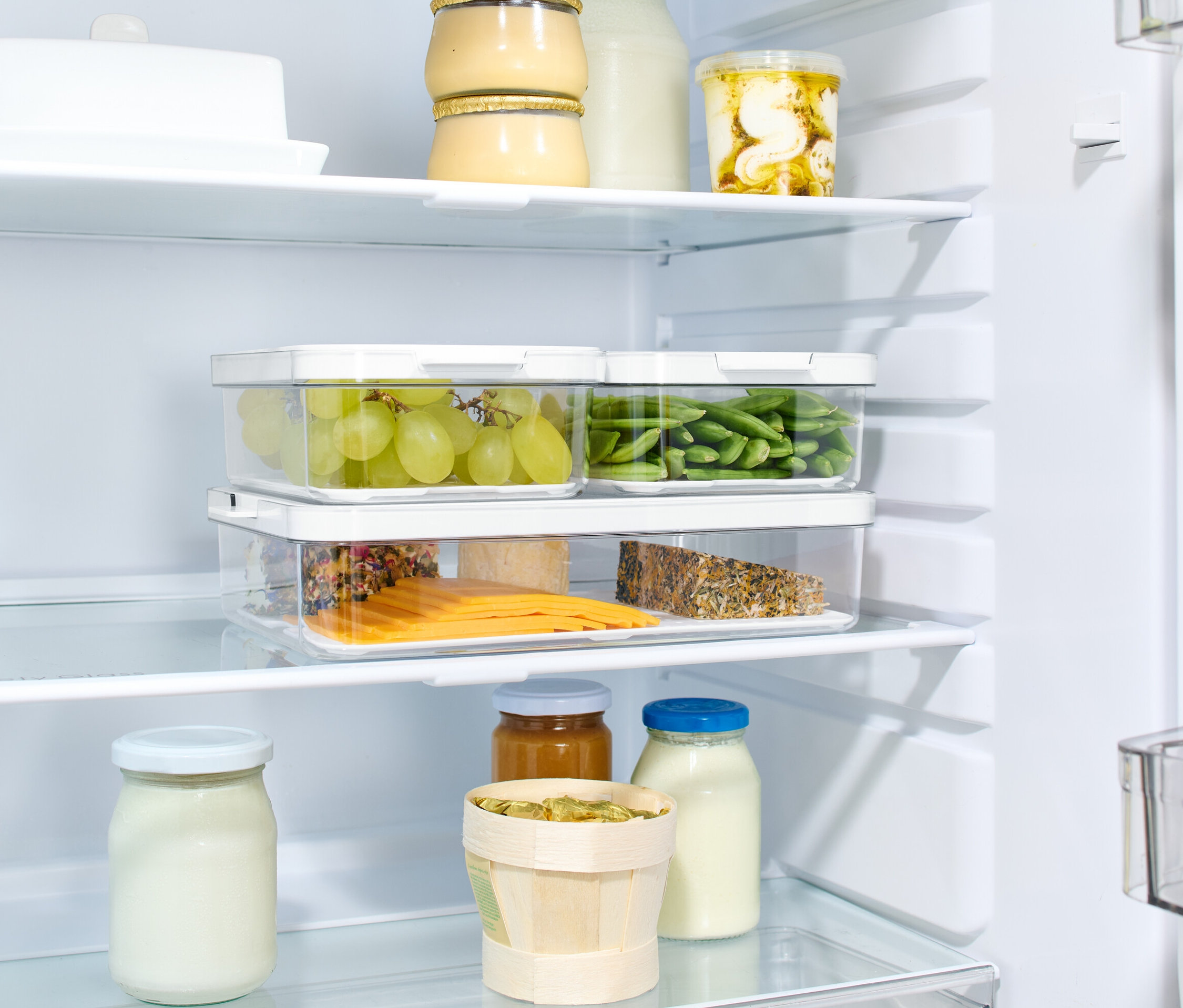 W lodówce znajdują się 3 pojemniki do przechowywania z pokrywkami, wypełnione zielonymi winogronami, zieloną fasolką szparagową i plasterkami sera.