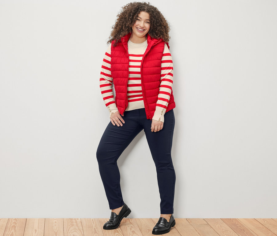 Kobieta stoi i uśmiecha się do kamery, ma na sobie czerwony sweter w paski, czerwoną kamizelkę pikowaną z kapturem i granatowe spodnie z bengaliny.