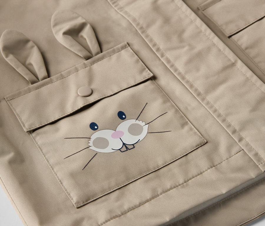 Zbliżenie na beżową kurtkę przeciwdeszczową dla dzieci z kieszenią z motywem króliczka.