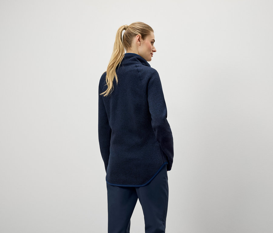 Z tyłu widać kobietę z blond włosami związanymi w koński ogon, ubraną w granatowy sweter i spodnie.