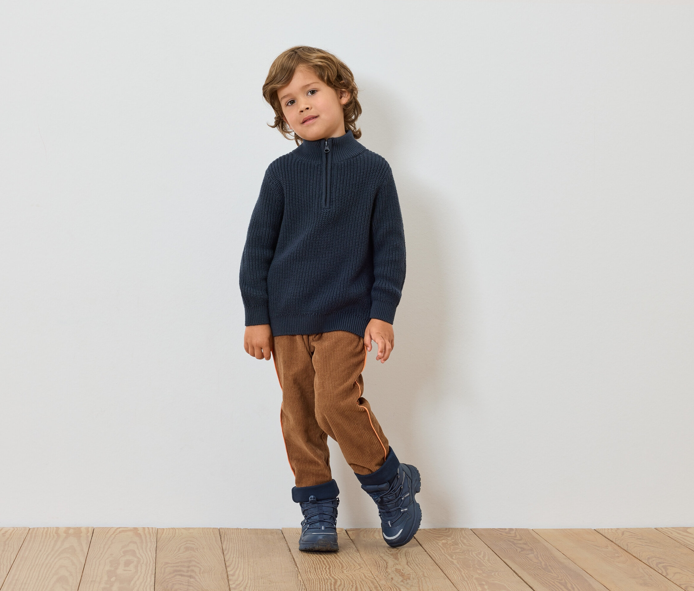 Chłopiec stoi przed białą ścianą. Ma na sobie granatowy sweter, brązowe spodnie i niebieskie buty.