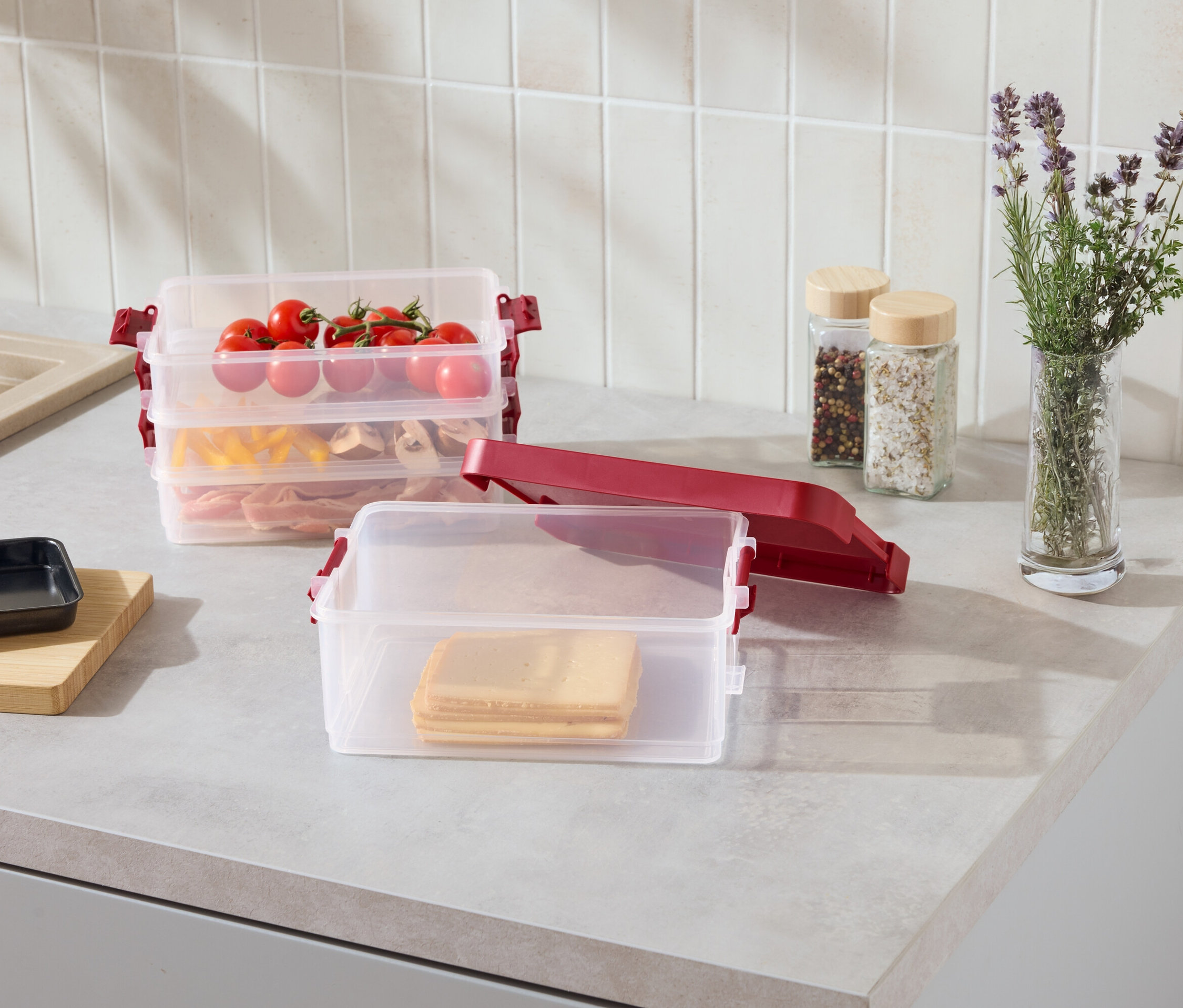 Na blacie kuchennym stoją przezroczyste, ustawione jeden na drugim pojemniki z czerwonymi klipsami, wypełnione plastrami sera, makaronem, mięsem i pomidorami.