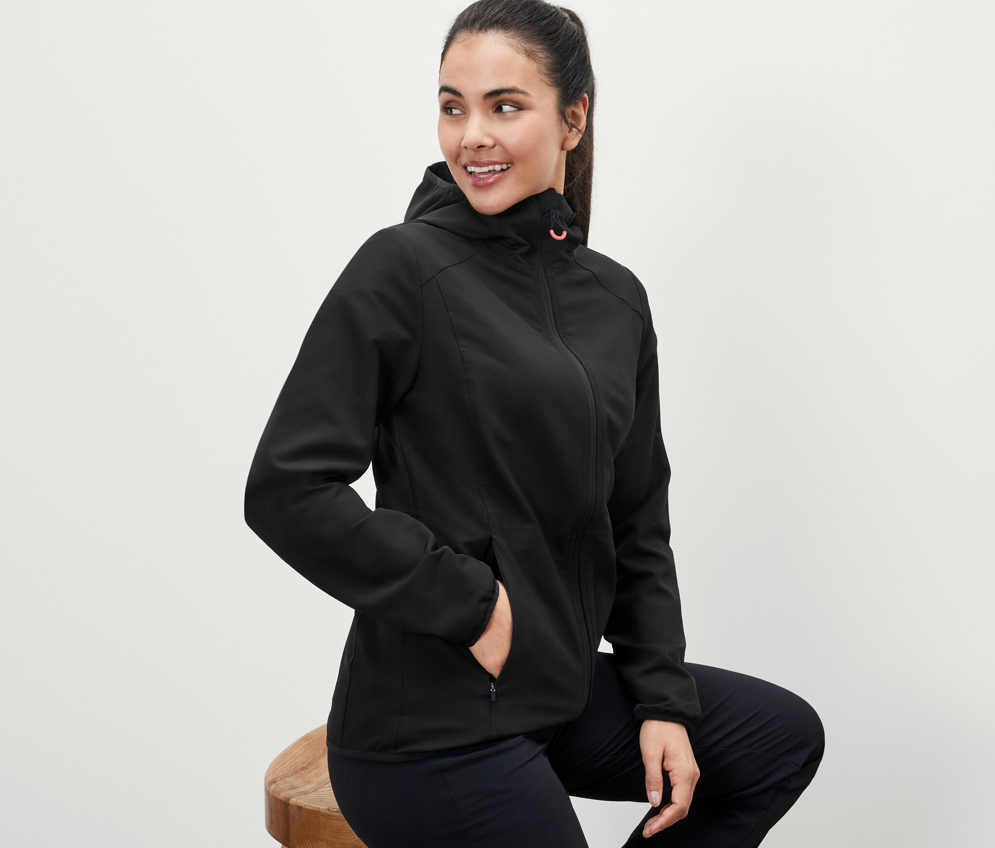 Kobieta siedzi na stołku i ma na sobie czarną kurtkę softshellową.