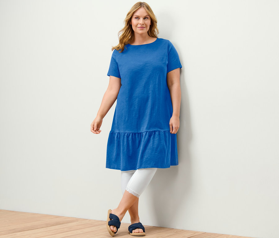Kobieta pozuje w kobaltowej sukience dżersejowej z falbaną, białych legginsach i niebieskich sandałach.