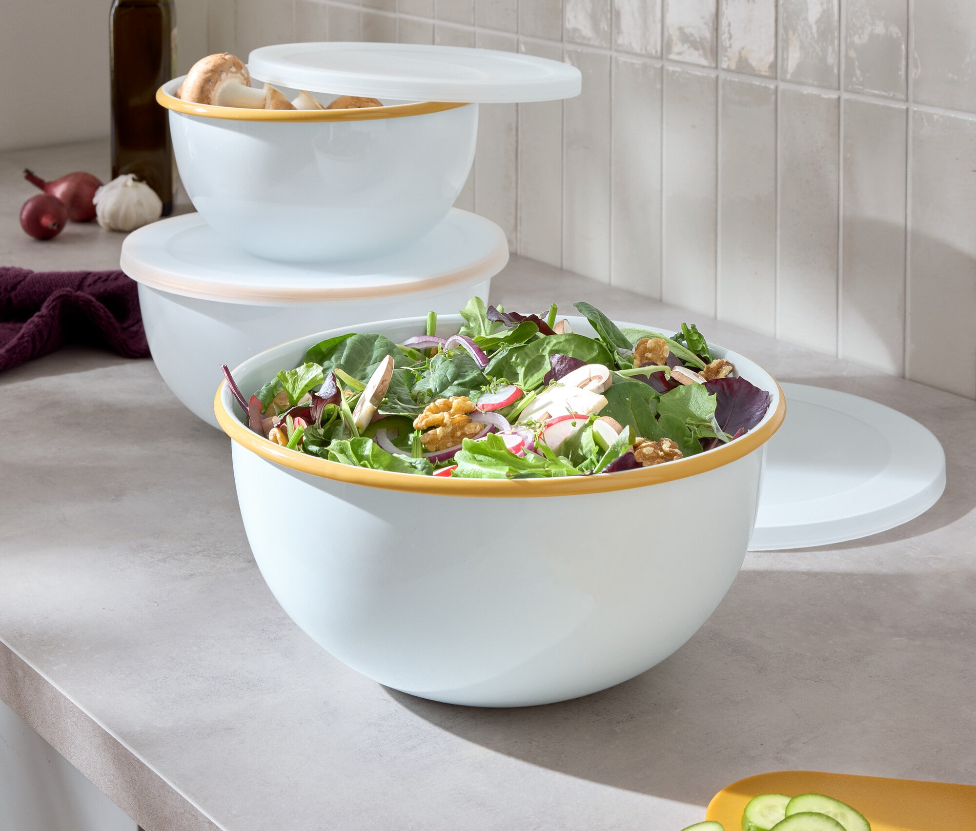 Trzy białe miski z kolorowym brzegiem ułożone jedna na drugiej, największa miska zawiera sałatę, obok leży żółta deska do krojenia z pokrojonym ogórkiem.