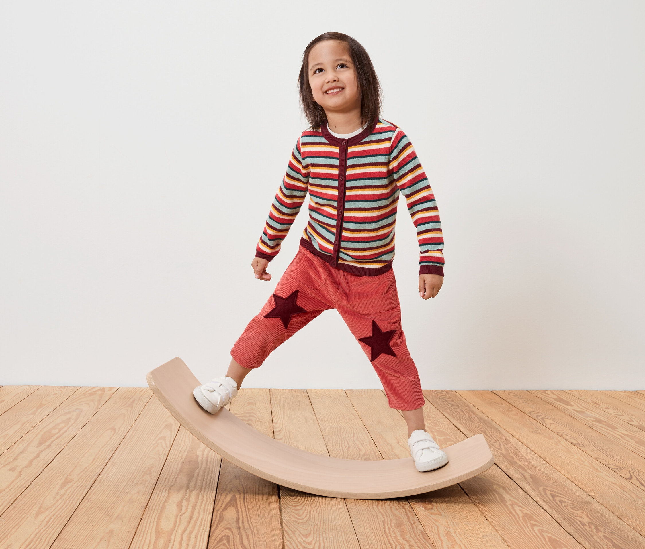 Dziecko stoi na drewnianej desce do balansowania, ma na sobie sweter w paski i spodnie z gwiazdami.