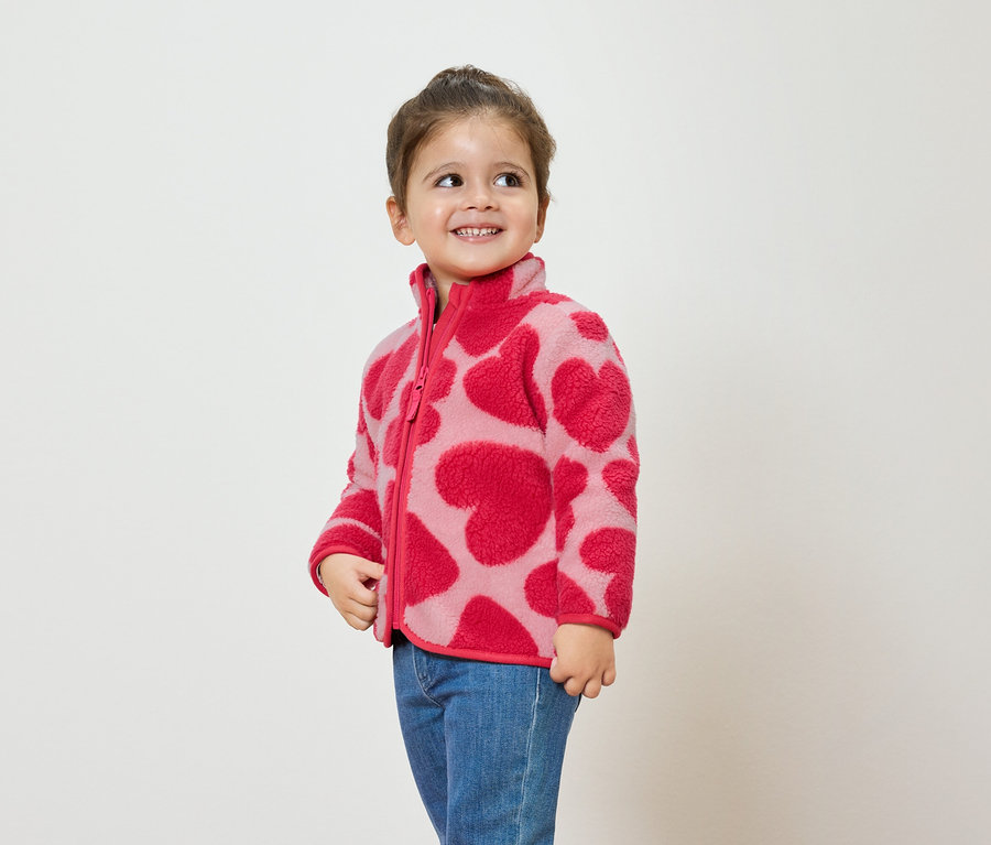 Dziewczynka w różowej bluzie polarowej w serca i niebieskich dżinsach uśmiecha się i patrzy w bok.