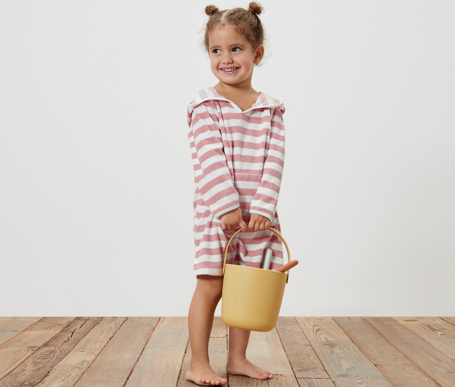 Uśmiechnięta dziewczynka trzyma żółte wiaderko i ma na sobie ponczo z tkaniny frotté w różowe paski.