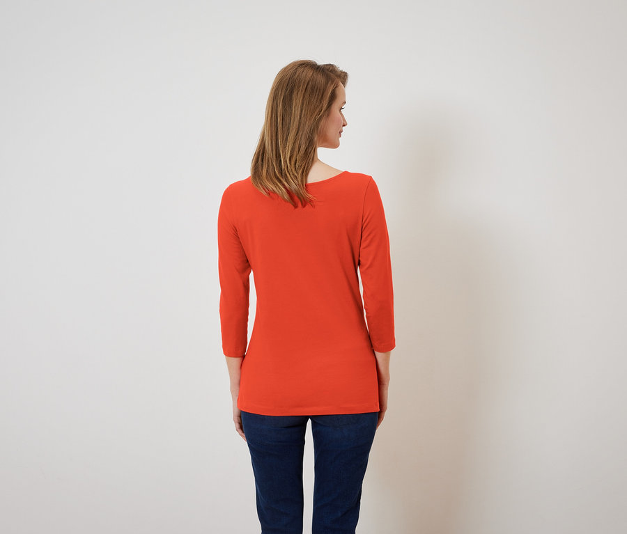 Kobieta tyłem do kamery ma na sobie pomarańczową koszulkę z rękawami trzy czwarte i granatowe dżinsy.